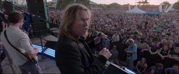 Movie still from “Song to Song” (2017), directed by Terrence Malick – A man with long blonde hair is holding a microphone in front of a crowd; Medium shot, Over the shoulder angle