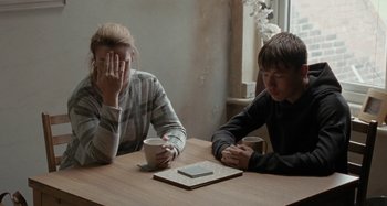 Movie still from “Sorry We Missed You” (2019), directed by Ken Loach – Two people sitting at a table with a cup of coffee; Medium shot, Over the shoulder angle