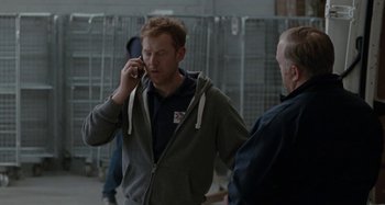 Movie still from “Sorry We Missed You” (2019), directed by Ken Loach – A man talking on a cell phone while standing next to another man; Medium shot, Over the shoulder angle