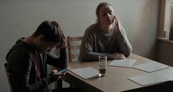 Movie still from “Sorry We Missed You” (2019), directed by Ken Loach – Two people sitting at a wooden table with a cell phone; Medium shot, Over the shoulder angle