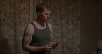 Movie still from “Sorry We Missed You” (2019), directed by Ken Loach – A man in a tank top looking at his cell phone; Medium shot, Over the shoulder angle