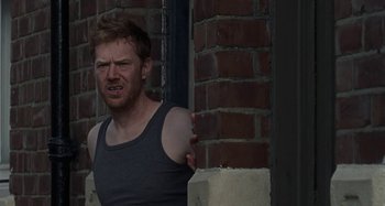 Movie still from “Sorry We Missed You” (2019), directed by Ken Loach – A man in a black tank top is leaning against a brick wall; Medium shot, Over the shoulder angle