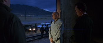 Movie still from “Species” (1995), directed by Roger Donaldson – A man in a trench coat standing next to a train track; Medium shot, Over the shoulder angle