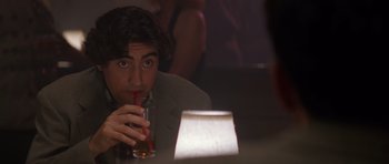 Movie still from “Species” (1995), directed by Roger Donaldson – A man sitting at a table drinking a beverage; Close Up shot, Over the shoulder angle