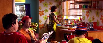 Movie still from “Speed Racer” (2008), directed by Lilly Wachowski – A woman in a kitchen reading a magazine while another woman stands at the sink; Medium shot, Over the shoulder angle