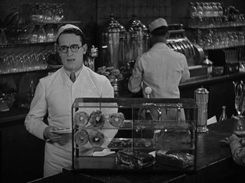 Movie still from “Speedy” (1928), directed by Ted Wilde – A black and white photo of a man holding a tray of donuts; Medium shot, Over the shoulder angle