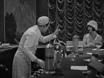 Movie still from “Speedy” (1928), directed by Ted Wilde – A man standing at a dining table with another man sitting at a table; Wide shot, High angle