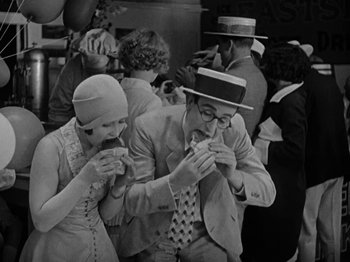 Movie still from “Speedy” (1928), directed by Ted Wilde – A man and a woman eating hot dogs at an event; Medium shot, High angle