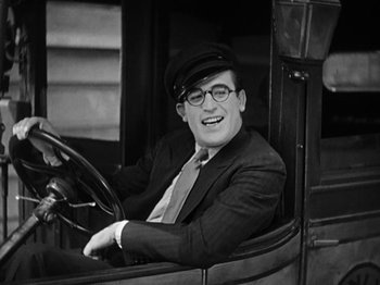 Movie still from “Speedy” (1928), directed by Ted Wilde – A man wearing a suit and hat sitting in the driver's seat of an automobile; Close Up shot, Low angle