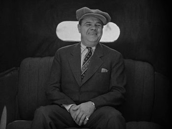 Movie still from “Speedy” (1928), directed by Ted Wilde – A man sitting on a couch wearing a suit and a hat; Medium shot, Low angle