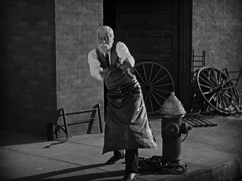 Movie still from “Speedy” (1928), directed by Ted Wilde – An old man standing next to a fire hydrant on the side of the street; Wide shot, Low angle