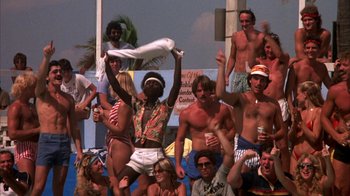 Movie still from “Spring Break” (1983), directed by Sean S. Cunningham – A group of people standing in front of a crowd; Wide shot, High angle