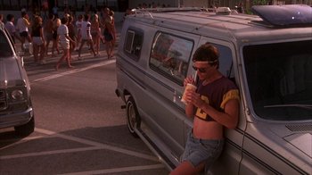 Movie still from “Spring Break” (1983), directed by Sean S. Cunningham – A man sitting on the side of a van eating a hot dog; Wide shot, Low angle