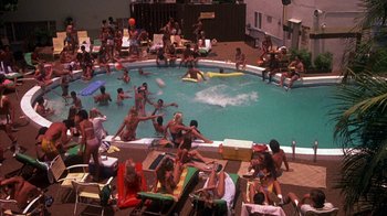 Movie still from “Spring Break” (1983), directed by Sean S. Cunningham – A crowd of people sitting and standing around a swimming pool; Extreme Wide shot, High angle