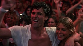 Movie still from “Spring Break” (1983), directed by Sean S. Cunningham – A man and a woman smiling for the camera in front of a crowd; Close Up shot, Low angle