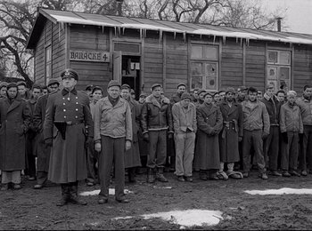 Movie still from “Stalag 17” (1953), directed by Billy Wilder – A group of men standing next to each other in front of a building; Wide shot, Over the shoulder angle