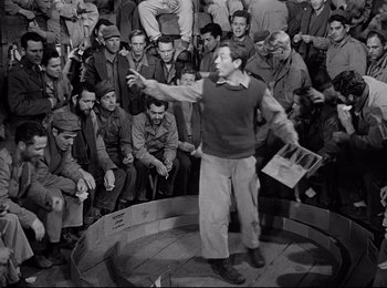 Movie still from “Stalag 17” (1953), directed by Billy Wilder – A man is standing in front of a group of men; Wide shot, High angle