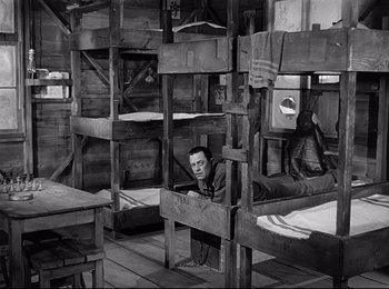 Movie still from “Stalag 17” (1953), directed by Billy Wilder – A man sitting on top of a bed in a room; Wide shot, High angle