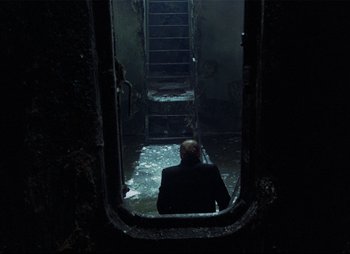 Movie still from “Stalker” (1979), directed by Andrei Tarkovsky – A man sitting in a doorway looking into a dark room; Wide shot, High angle