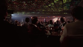 Movie still from “Star 80” (1983), directed by Bob Fosse – A group of people sitting at tables in front of a microphone; Extreme Wide shot, High angle