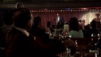 Movie still from “Star 80” (1983), directed by Bob Fosse – A group of people sitting at a table with a microphone in front of them; Wide shot, Over the shoulder angle