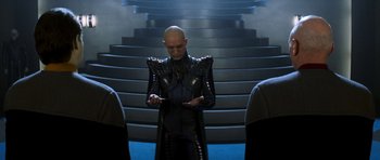 Movie still from “Star Trek: Nemesis” (2002), directed by Stuart Baird – A bald man in a black leather jacket is looking at his cell phone in front of some stairs; Medium shot, Over the shoulder angle