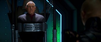Movie still from “Star Trek: Nemesis” (2002), directed by Stuart Baird – An older man in a red shirt is standing in front of a green wall; Medium shot, Low angle