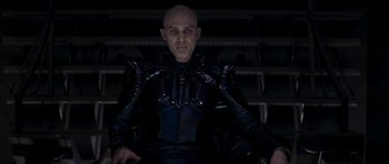 Movie still from “Star Trek: Nemesis” (2002), directed by Stuart Baird – A bald man in a leather jacket sitting in a chair in front of stairs; Medium shot, Low angle