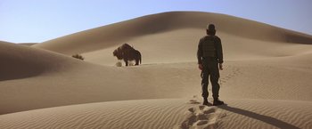 Movie still from “Stargate” (1994), directed by Roland Emmerich – A man standing on top of a sand dune next to an animal; Wide shot, Low angle