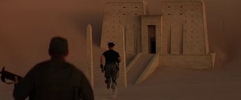 Movie still from “Stargate” (1994), directed by Roland Emmerich – A man walking down a dirt road in front of a building; Wide shot, Over the shoulder angle