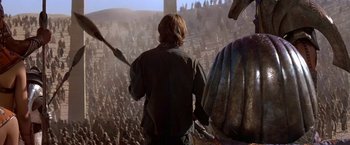 Movie still from “Stargate” (1994), directed by Roland Emmerich – A man standing in front of an audience holding a spear; Wide shot, High angle