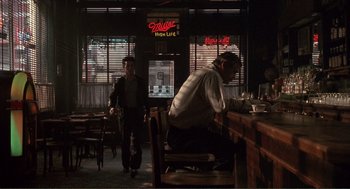 Movie still from “State of Grace” (1990), directed by Phil Joanou – A man sitting at a table in front of a neon sign; Wide shot, Over the shoulder angle