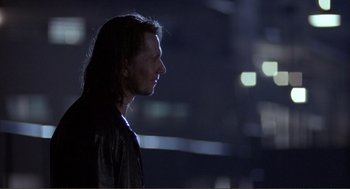 Movie still from “State of Grace” (1990), directed by Phil Joanou – A man with long black hair standing in the dark; Close Up shot, Over the shoulder angle