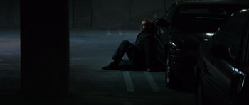 Movie still from “State of Play” (2009), directed by Kevin Macdonald – A man sitting on the ground in a parking lot next to a parked car; Wide shot, High angle
