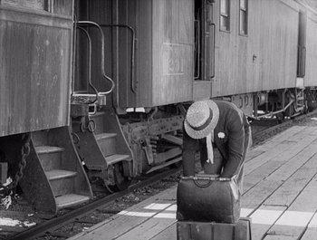 Movie still from “Steamboat Bill, Jr.” (1928), directed by Buster Keaton – An old photo of a man with a hat and a suit case; Wide shot, High angle