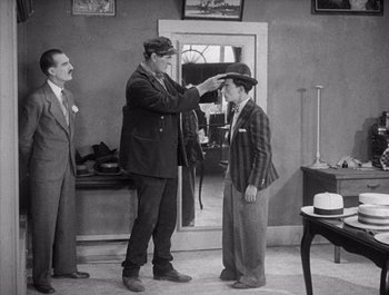 Movie still from “Steamboat Bill, Jr.” (1928), directed by Buster Keaton – A man in a suit and tie is putting a hat on a young man; Medium shot, Over the shoulder angle