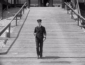 Movie still from “Steamboat Bill, Jr.” (1928), directed by Buster Keaton – A man in a uniform walking across a wooden bridge; Wide shot, High angle