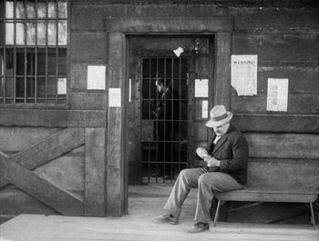 Movie still from “Steamboat Bill, Jr.” (1928), directed by Buster Keaton – An older man sitting on a bench in front of a building; Wide shot, High angle
