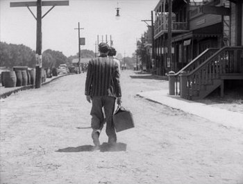 Movie still from “Steamboat Bill, Jr.” (1928), directed by Buster Keaton – A man walking down a street holding a briefcase; Wide shot, Over the shoulder angle