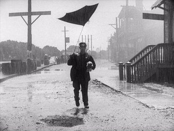 Movie still from “Steamboat Bill, Jr.” (1928), directed by Buster Keaton – A man holding an umbrella while walking down the street; Wide shot, Low angle