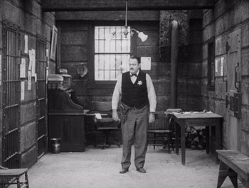 Movie still from “Steamboat Bill, Jr.” (1928), directed by Buster Keaton – A man standing in a room with a table and chair in front of him; Wide shot, High angle