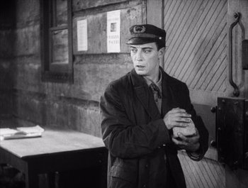 Movie still from “Steamboat Bill, Jr.” (1928), directed by Buster Keaton – A man wearing a hat and a jacket standing in front of a building; Medium shot, Low angle