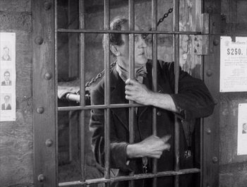 Movie still from “Steamboat Bill, Jr.” (1928), directed by Buster Keaton – A man standing behind a metal fence with chains on it; Medium shot, High angle