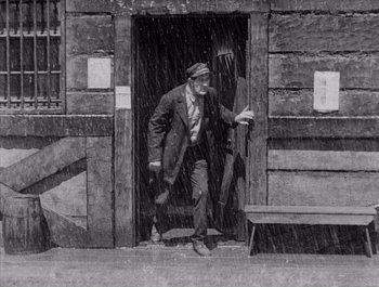 Movie still from “Steamboat Bill, Jr.” (1928), directed by Buster Keaton – An old photo of a man walking in the rain; Wide shot, High angle