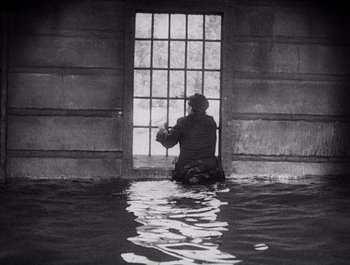 Movie still from “Steamboat Bill, Jr.” (1928), directed by Buster Keaton – A man sitting on the ground in front of a window; Wide shot, High angle