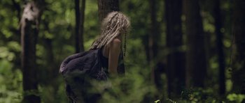 Movie still from “Still” (2018), directed by Takashi Doscher – A woman with dreads standing next to a tree in the woods; Medium shot, Low angle