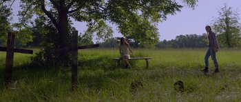 Movie still from “Still” (2018), directed by Takashi Doscher – A woman sitting on top of a wooden bench under a tree; Wide shot, High angle