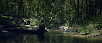 Movie still from “Still” (2018), directed by Takashi Doscher – A woman sitting on a tree stump in the middle of a river; Wide shot, High angle