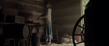 Movie still from “Still” (2018), directed by Takashi Doscher – A woman standing in front of a table holding a gun; Wide shot, High angle