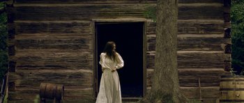 Movie still from “Still” (2018), directed by Takashi Doscher – A woman standing in front of an old log cabin; Wide shot, Low angle